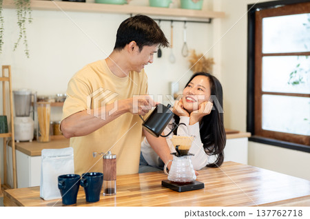 Asian woman smiling at man holding kettle pouring water in dripper making coffee at kitchen counter. 137762718