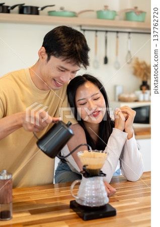 Asian woman leaning on man holding kettle pouring water in dripper making coffee at kitchen counter. Asian woman leaning on man holding kettle pouring water in dripper making coffee at kitchen counter. 137762720