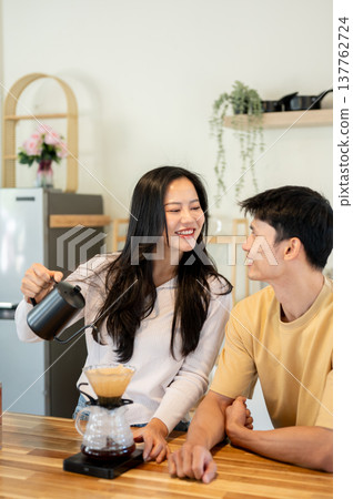 Asian man looking at a woman holding kettle pouring water making dripper coffee at kitchen counter. Asian man looking at a woman holding kettle pouring water making dripper coffee at kitchen counter. 137762724