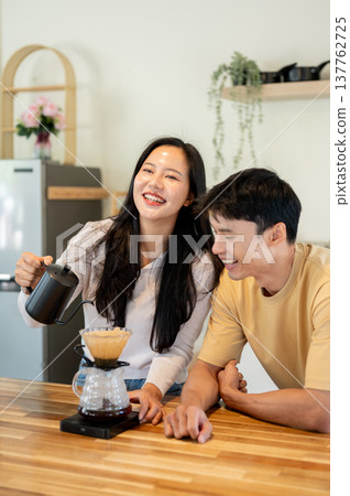 Laughing man looking at woman holding kettle pouring water making dripper coffee at kitchen counter. Laughing man looking at woman holding kettle pouring water making dripper coffee at kitchen counter. 137762725