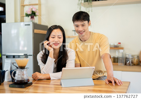 Asian man woman couple looking at tablet watching video while making drip coffee at kitchen counter. Asian man woman couple looking at tablet watching video while making drip coffee at kitchen counter. 137762727
