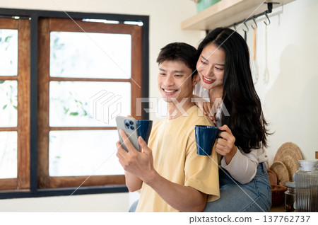 Happy asian woman and man couple holding coffee looking at phone sitting leaning on kitchen counter. 137762737