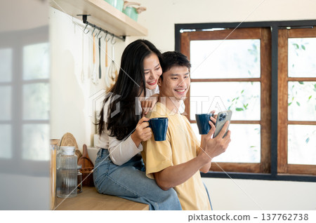 Young asian woman and man couple holding coffee looking at phone sitting leaning on kitchen counter. Young asian woman and man couple holding coffee looking at phone sitting leaning on kitchen counter. 137762738