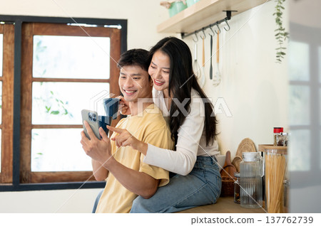 Asian woman and man couple holding coffee cup looking at phone sitting leaning on kitchen counter. Asian woman and man couple holding coffee cup looking at phone sitting leaning on kitchen counter. 137762739