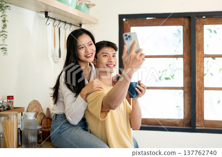Pretty asian woman and man couple holding coffee taking picture sitting leaning on kitchen counter. Pretty asian woman and man couple holding coffee taking picture sitting leaning on kitchen counter. 137762740
