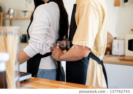 Close up of couple a man tying apron for woman girlfriend before cooking standing at kitchen counter Close up of couple a man tying apron for woman girlfriend before cooking standing at kitchen counter 137762743