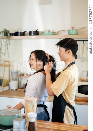 Asian couple a man tying woman girlfriend's hair before cooking food as standing at kitchen counter. Asian couple a man tying woman girlfriend's hair before cooking food as standing at kitchen counter. 137762744