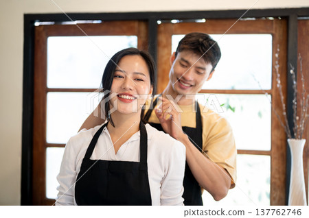 Asian couple a man tying woman girlfriend's hair before cooking preparing food standing in kitchen. Asian couple a man tying woman girlfriend's hair before cooking preparing food standing in kitchen. 137762746