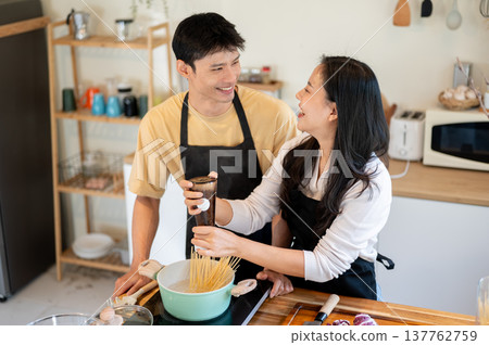 Asian couple a woman putting salt in boiling spaghetti aside smiling man at kitchen cooking counter. Asian couple a woman putting salt in boiling spaghetti aside smiling man at kitchen cooking counter. 137762759