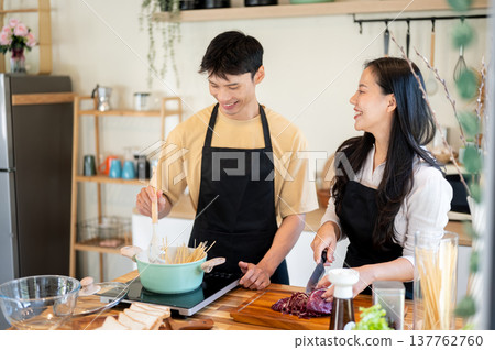Asian couple man holding spoon boiling spaghetti as woman girlfriend cuts veggie at kitchen counter. 137762760