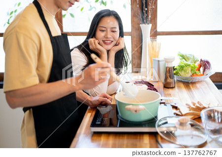 Asian woman girlfriend resting head on hands looking at boyfriend man cooking at kitchen counter. Asian woman girlfriend resting head on hands looking at boyfriend man cooking at kitchen counter. 137762767