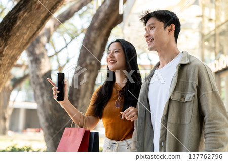 Asian couple woman holding shopping bag and phone pointing as linking arms walking with man in park. Asian couple woman holding shopping bag and phone pointing as linking arms walking with man in park. 137762796