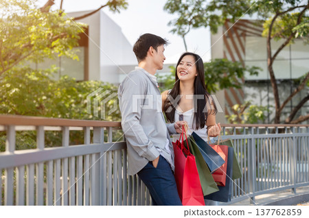 Asian couple a man holding shopping bags talking to a smiling woman while standing on bridge in park Asian couple a man holding shopping bags talking to a smiling woman while standing on bridge in park 137762859