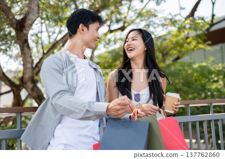 Asian couple a man talking to woman holding shopping bag and coffee while standing on bridge in park Asian couple a man talking to woman holding shopping bag and coffee while standing on bridge in park 137762860