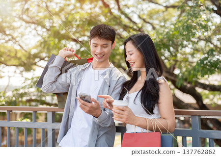 Asian couple a man holding shopping bag and phone showing to woman while standing on bridge in park. Asian couple a man holding shopping bag and phone showing to woman while standing on bridge in park. 137762862