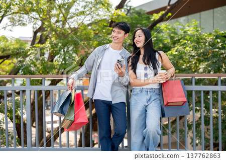 Asian couple man carrying shopping bag and phone woman holding coffee standing on footbridge in park Asian couple man carrying shopping bag and phone woman holding coffee standing on footbridge in park 137762863