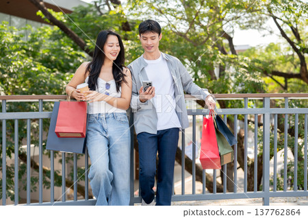 Asian couple a man showing phone to woman holding coffee and shopping bag leaning on railing in park Asian couple a man showing phone to woman holding coffee and shopping bag leaning on railing in park 137762864
