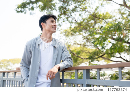 Smart asian man standing with elbow on railing looking around a footbridge aside green tree in park. Smart asian man standing with elbow on railing looking around a footbridge aside green tree in park. 137762874