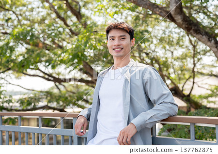 Smiling asian man standing with elbows on railing leaning against footbridge aside a tree in park. Smiling asian man standing with elbows on railing leaning against footbridge aside a tree in park. 137762875