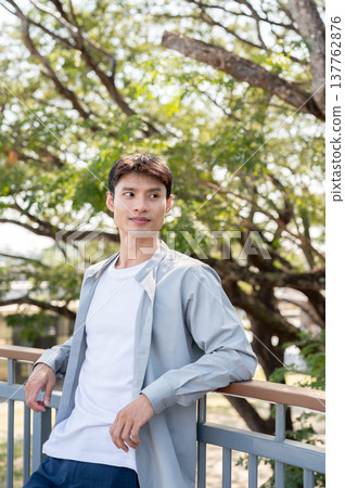 Handsome asian man standing with elbows on railing leaning against footbridge aside a tree in park. 137762876