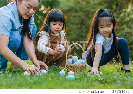 mother and her two daughters playing and hunting for Easter eggs into a basket on green grass in garden mother and her two daughters playing and hunting for Easter eggs into a basket on green grass in garden 137763052