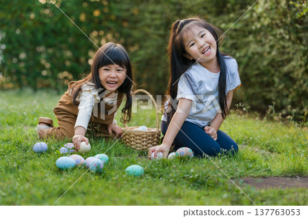 two happy child girl playing together and hunting for Easter eggs into basket on green grass in garden two happy child girl playing together and hunting for Easter eggs into basket on green grass in garden 137763053