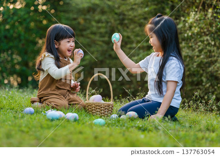 two happy child girl playing together and hunting for Easter eggs into basket on green grass in garden two happy child girl playing together and hunting for Easter eggs into basket on green grass in garden 137763054