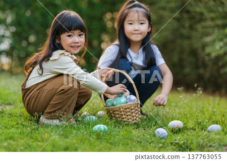 two happy child girl playing together and hunting for Easter eggs into basket on green grass in garden 137763055