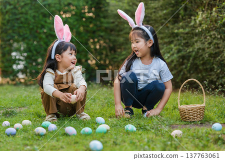 two happy child girl wearing bunny ears playing together and hunting for Easter eggs into basket on green grass in garden two happy child girl wearing bunny ears playing together and hunting for Easter eggs into basket on green grass in garden 137763061