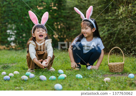 two happy child girl wearing bunny ears playing together and hunting for Easter eggs into basket on green grass in garden two happy child girl wearing bunny ears playing together and hunting for Easter eggs into basket on green grass in garden 137763062