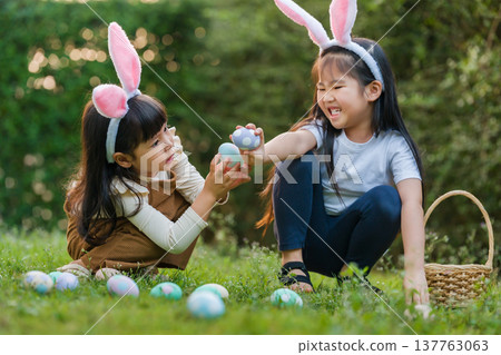 two happy child girl wearing bunny ears playing together and hunting for Easter eggs into basket on green grass in garden two happy child girl wearing bunny ears playing together and hunting for Easter eggs into basket on green grass in garden 137763063