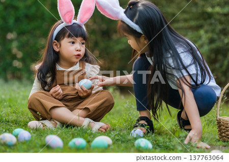 two child girl wearing bunny ears playing together and hunting for Easter eggs into basket on green grass in garden two child girl wearing bunny ears playing together and hunting for Easter eggs into basket on green grass in garden 137763064