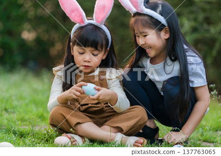 two happy child girl wearing bunny ears playing together and hunting for Easter eggs into basket on green grass in garden two happy child girl wearing bunny ears playing together and hunting for Easter eggs into basket on green grass in garden 137763065
