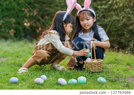 two child girl wearing bunny ears playing together and hunting for Easter eggs into basket on green grass in garden two child girl wearing bunny ears playing together and hunting for Easter eggs into basket on green grass in garden 137763066