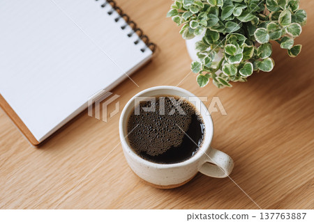 An overhead photo of a natural workspace with a coffee cup, a notebook, and a houseplant. 137763887