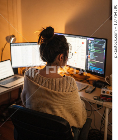 Person working late at computer desk with multiple screens, cozy sweater, and warm lighting creating focused and comfortable atmosphere 137764590
