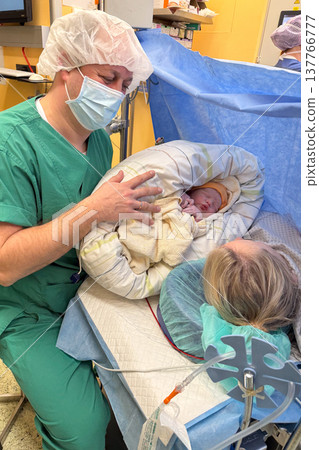 Father holding newborn baby girl in operating room while mother lies on surgical table during cesarean section. Emotional moment of first contact between parents and newborn in hospital. Father holding newborn baby girl in operating room while mother lies on surgical table during cesarean section. Emotional moment of first contact between parents and newborn in hospital. 137766777