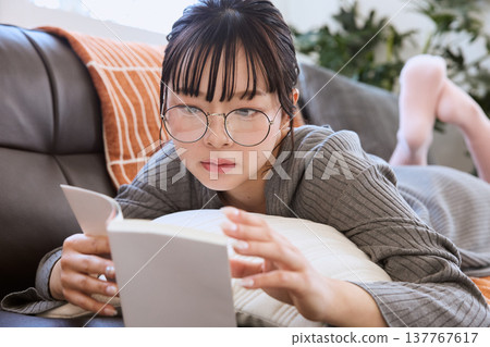 一位戴眼鏡的女士正在看書;她在家享受閱讀時光。 一位戴眼鏡的女士正在看書;她在家享受閱讀時光。 137767617