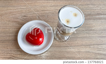 Elegant red heart mirror glaze mousse cake served with layered latte macchiato on a wooden table Elegant red heart mirror glaze mousse cake served with layered latte macchiato on a wooden table 137768021