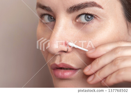 Inflamed nose piercing. Caucasian woman swabbing the piercing site with chlorhexidine on a cotton swab. Post piercing care. Close up photo 137768502