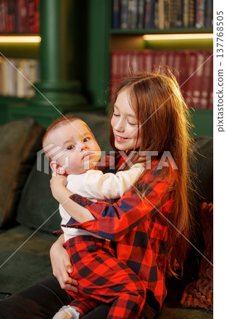 Little girl holds baby brother in cozy room with books Little girl holds baby brother in cozy room with books 137768505