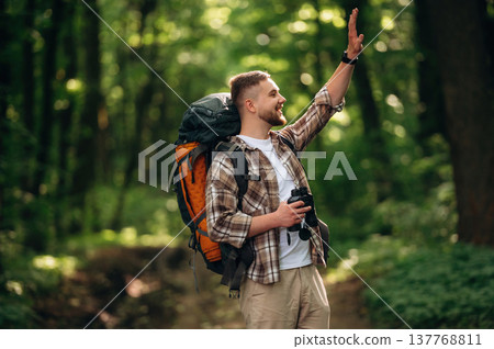 Side view, standing, holding binoculars. Male tourist is with backpack in the forest Side view, standing, holding binoculars. Male tourist is with backpack in the forest 137768811