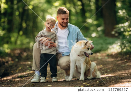 Positive facial expression, sitting. Father with son and dog are in the forest Positive facial expression, sitting. Father with son and dog are in the forest 137769237