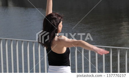 A woman in a black tank top stretches on the waterfront, enjoying movement and sunlight. This shot captures the harmony between body and nature, emphasizing the importance of an active lifestyle. A woman in a black tank top stretches on the waterfront, enjoying movement and sunlight. This shot captures the harmony between body and nature, emphasizing the importance of an active lifestyle. 137770237