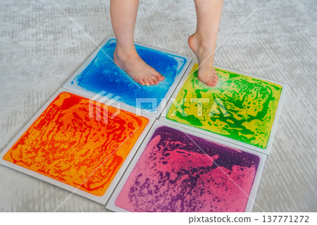 Child feet standing on colorful sensory gel mat on floor during tactile play activity. Sensory stimulation, autism support and occupational therapy development concept. Child feet standing on colorful sensory gel mat on floor during tactile play activity. Sensory stimulation, autism support and occupational therapy development concept. 137771272