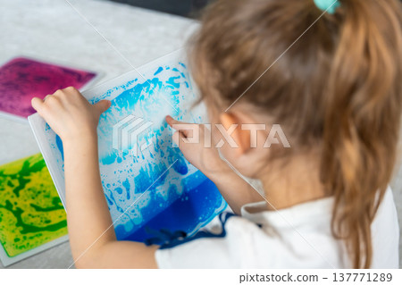 Little girl holding colorful sensory gel mat and pressing surface with finger while drawing shapes. Tactile stimulation and sensory development concept for autism support and occupational therapy. Little girl holding colorful sensory gel mat and pressing surface with finger while drawing shapes. Tactile stimulation and sensory development concept for autism support and occupational therapy. 137771289
