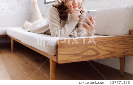 一名女子麵朝下躺在沙發上,正在使用智慧型手機。 一名女子麵朝下躺在沙發上,正在使用智慧型手機。 137771326