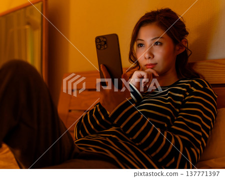 一位女士躺在床上看著她的智慧型手機，鏡頭捕捉到了她的側臉。 137771397