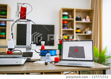 A 3D printer is actively creating a red and white object while a laptop displays the design on its screen, surrounded by filament spools and supplies A 3D printer is actively creating a red and white object while a laptop displays the design on its screen, surrounded by filament spools and supplies 137771552