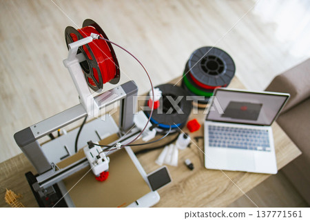 A 3D printer is set up on a wooden table with spools of filament and a laptop displaying a 3D model A 3D printer is set up on a wooden table with spools of filament and a laptop displaying a 3D model 137771561